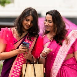 Two Indian women in traditional attire looking at a smartphone, representing diverse financial choices